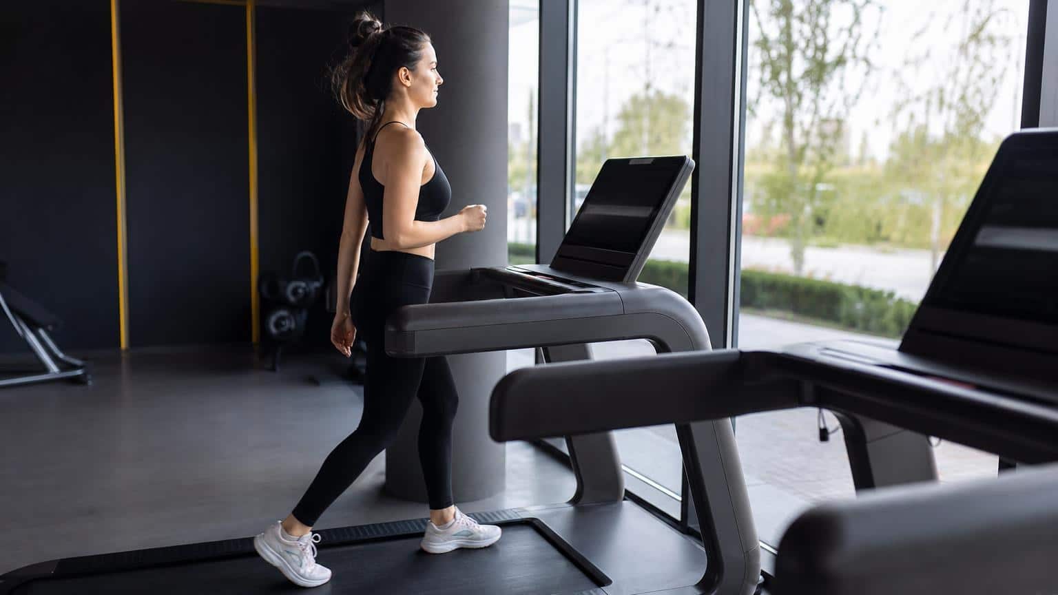 Woman in black athletic wear walking on a treadmill in a modern gym facing a large window with a view of green trees. Woman in black athletic wear walking on a treadmill in a modern gym facing a large window with a view of green trees.